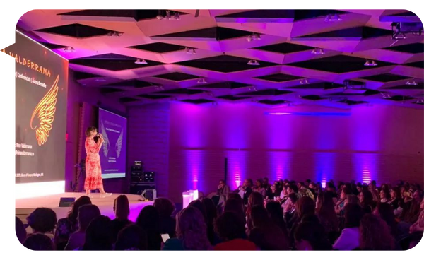 Nina Valderrama dando una conferencia en un auditorio iluminado con luces púrpuras, frente a una audiencia numerosa.