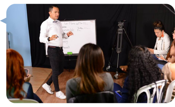 Leo Parra impartiendo una conferencia frente a un grupo de personas, utilizando una pizarra blanca como apoyo visual y destacando con su distintivo corbatín.