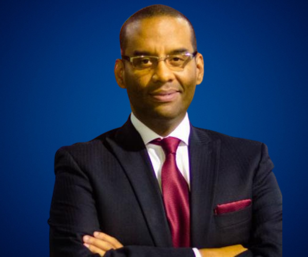 Fotografía de Hermes Ruiz, vestido con traje oscuro y corbata burdeos, posando con los brazos cruzados sobre un fondo azul profesional.