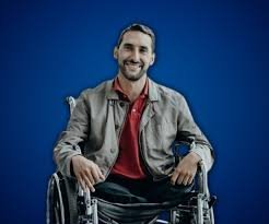 Fotografía de Juan Manuel Montañez, sonriendo mientras posa en su silla de ruedas con chaqueta gris y camiseta roja, sobre un fondo azul liso.