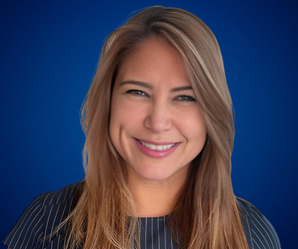 Fotografía de Linda De La Torre, sonriente y de frente, sobre un fondo azul profesional. Luce una blusa oscura con líneas claras, cabello suelto y expresión amigable.
