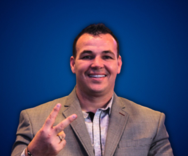 Fotografía de Alexis "El Pulpo" Viera, sonriente, haciendo el gesto de paz con los dedos, vestido con saco gris claro sobre camisa, posando frente a un fondo azul sólido.