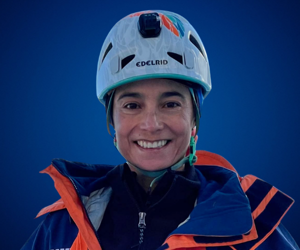 Ana María Giraldo, conferencista motivacional colombiana, usando casco de montaña y chaqueta técnica, sonriente sobre fondo azul