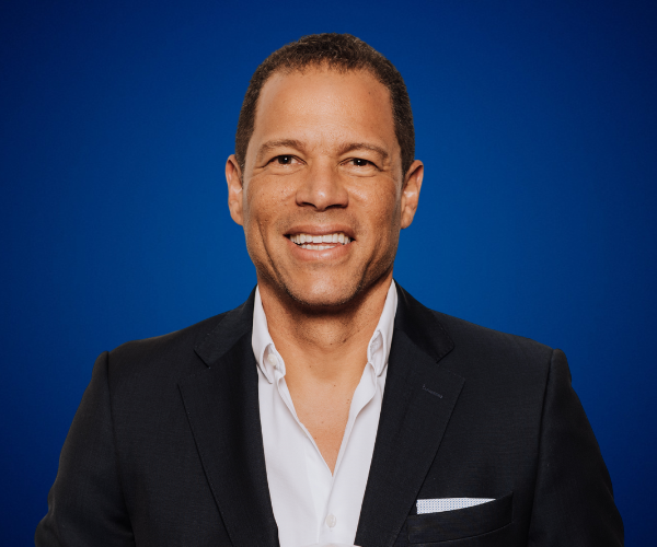 Óscar Córdoba, exfutbolista y conferencista motivacional colombiano, sonriente con traje formal sobre fondo azul