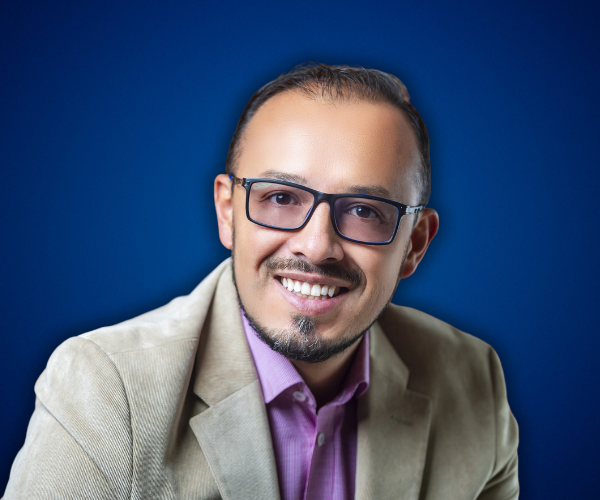 Fotografía de Oscar Rodríguez Lemus, retrato profesional de medio cuerpo con fondo azul, luciendo saco beige y camisa lila, con expresión sonriente y gafas oscuras.