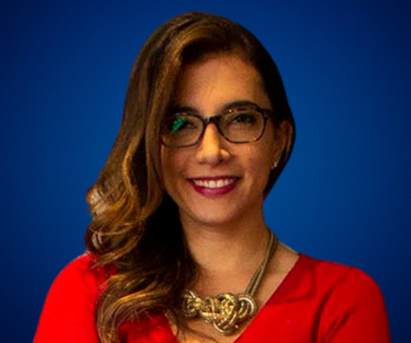 Fotografía de Pilar Ibañez, mujer sonriente con gafas y cabello ondulado, vestida con blusa roja y collar dorado, sobre fondo azul liso.